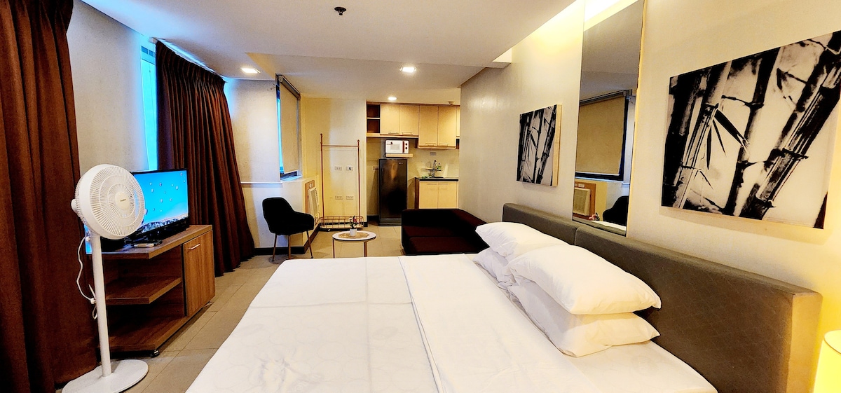 A spacious sleeping area features a king size bed with white linens and a neutral-toned headboard. Natural light enters through the windows adorned with dark curtains. A kitchenette is visible in the background, complemented by a dining area. A television and a fan are also present in the room.