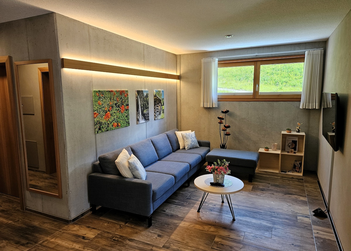 A modern living area features a sectional sofa adorned with soft cushions, positioned next to a small table. Natural light filters in through a large window, highlighting the subtle green landscape outside. Wall art adds color to the neutral walls, enhancing the inviting ambiance.