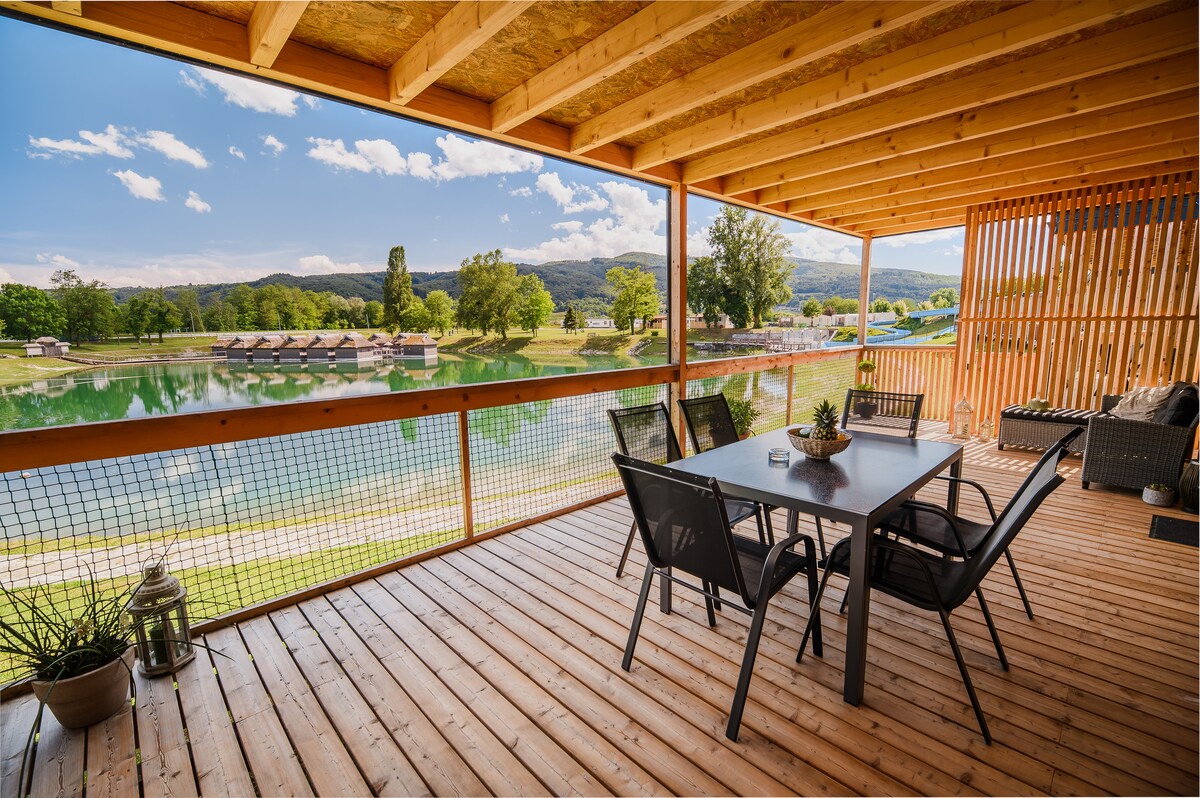 A spacious terrace is surrounded by wooden beams and features a dining table set with several chairs. The area overlooks a serene lake, with greenery extending in the background under a clear blue sky, creating a peaceful outdoor space.