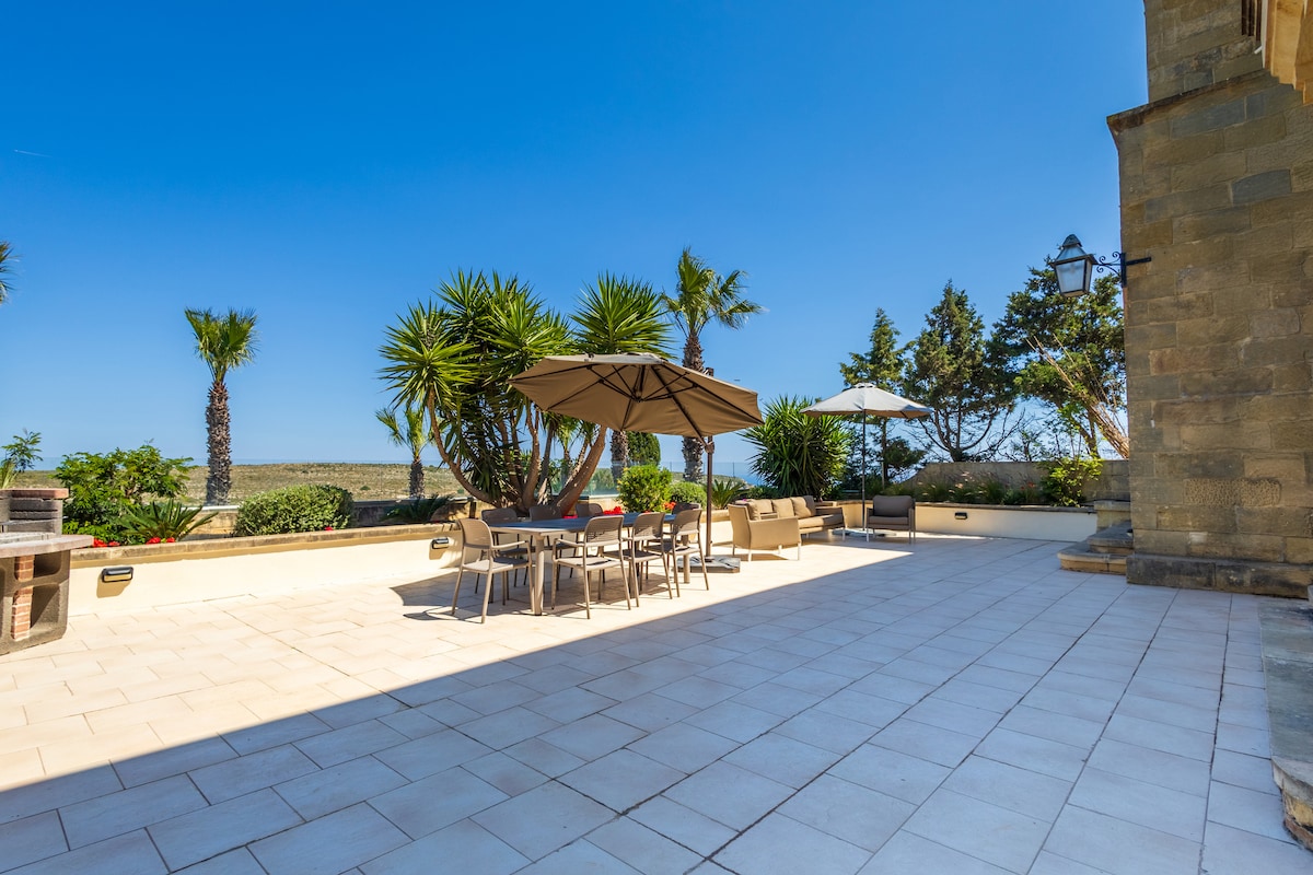 An expansive outdoor terrace is presented, featuring a large umbrella over a dining table surrounded by several chairs. Lush palm trees and shrubs provide greenery in the background, while the clear blue sky enhances the serene atmosphere of the space.