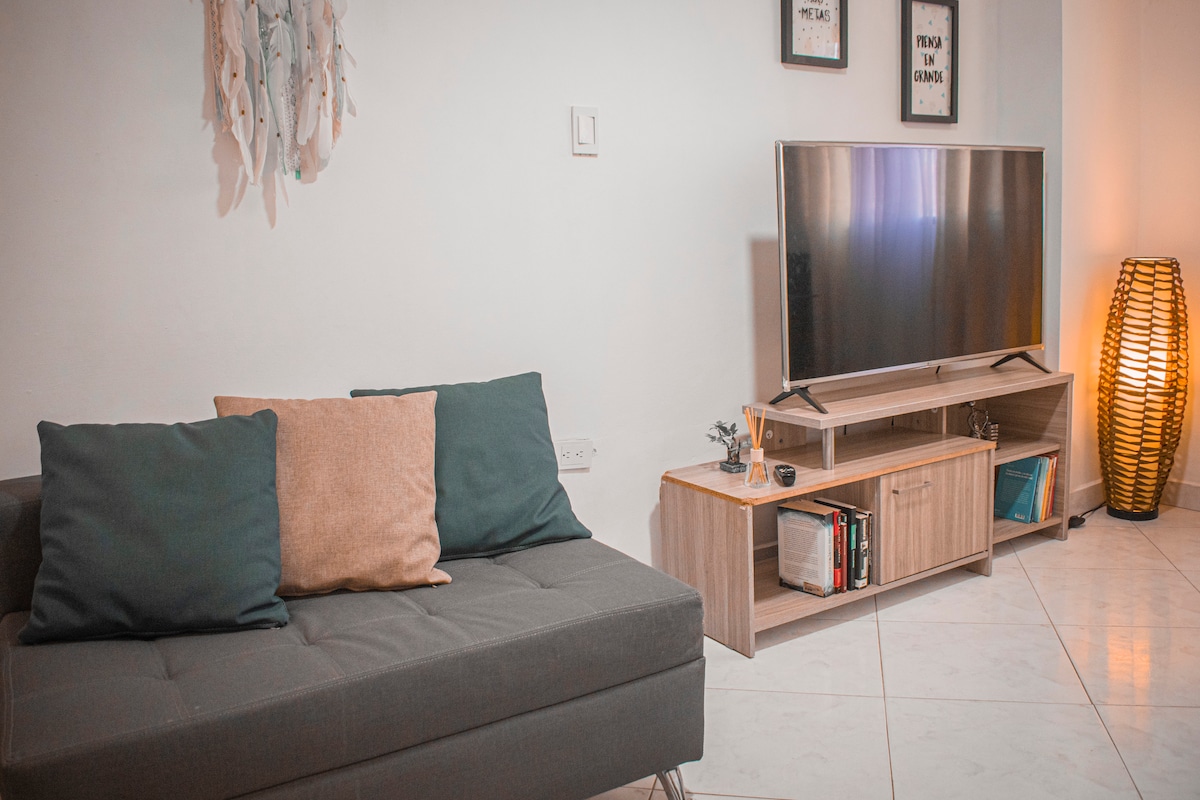 A modern living area features a dark gray sofa with decorative pillows, positioned in front of a flat-screen television. A stylish wooden console holds books and decorative items. Soft lighting is provided by a woven lamp, creating a calm and comfortable environment.
