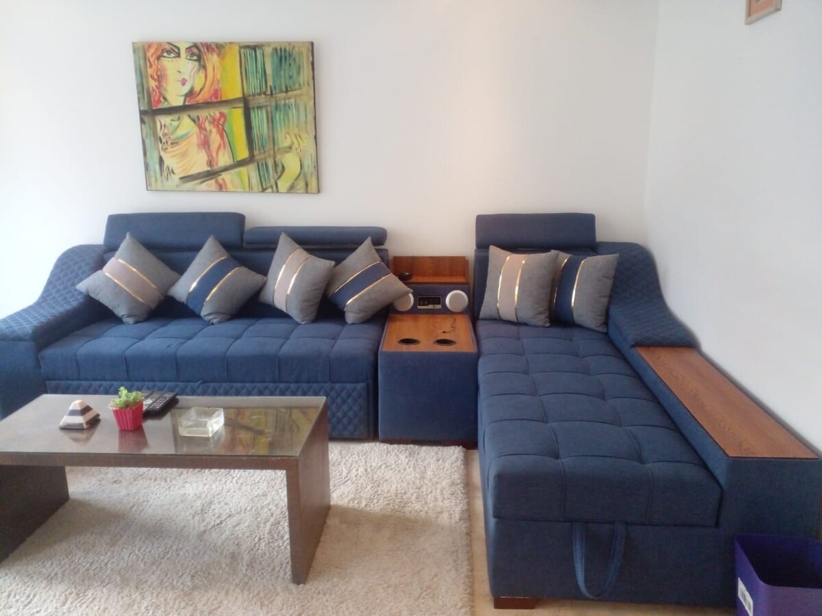 A spacious living area features a large, blue sectional sofa adorned with decorative cushions. A wooden side table is situated next to the sofa, and a small plant adds a touch of greenery. The light carpet enhances the room's relaxed ambiance.