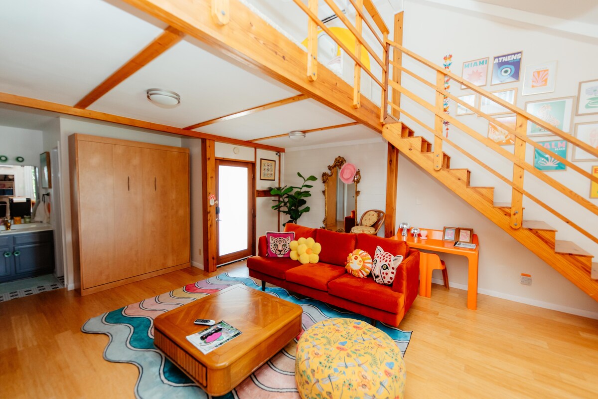 A vibrant living area showcases a bright orange sofa adorned with decorative pillows. Underfoot, a multi-colored area rug adds warmth. A wooden coffee table sits at the center, and a staircase with wooden railings leads to the loft. Greenery complements the colorful wall decor.