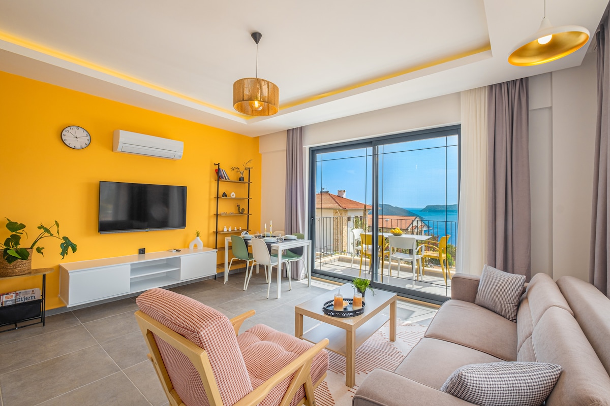 A bright living area features a large window with views of the sea and Megisti. An entertainment unit with a television is positioned on a light grey wall, while a comfortable sofa and armchair provide seating. A dining table is set against the backdrop of a vibrant orange wall.