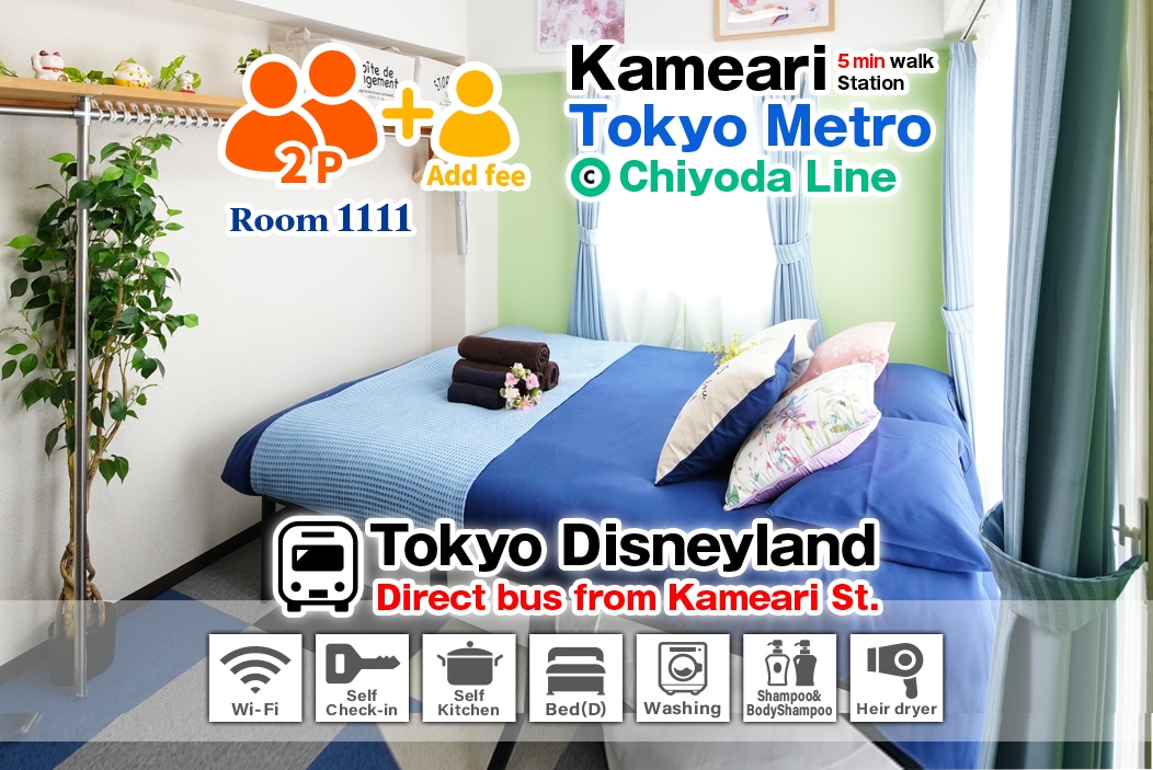 A compact room features a spacious double bed with decorative pillows and a cozy sofa bed adjacent. Brightening the space, large windows showcase natural light, while a neatly organized kitchenette and bathroom amenities are visible in the background.