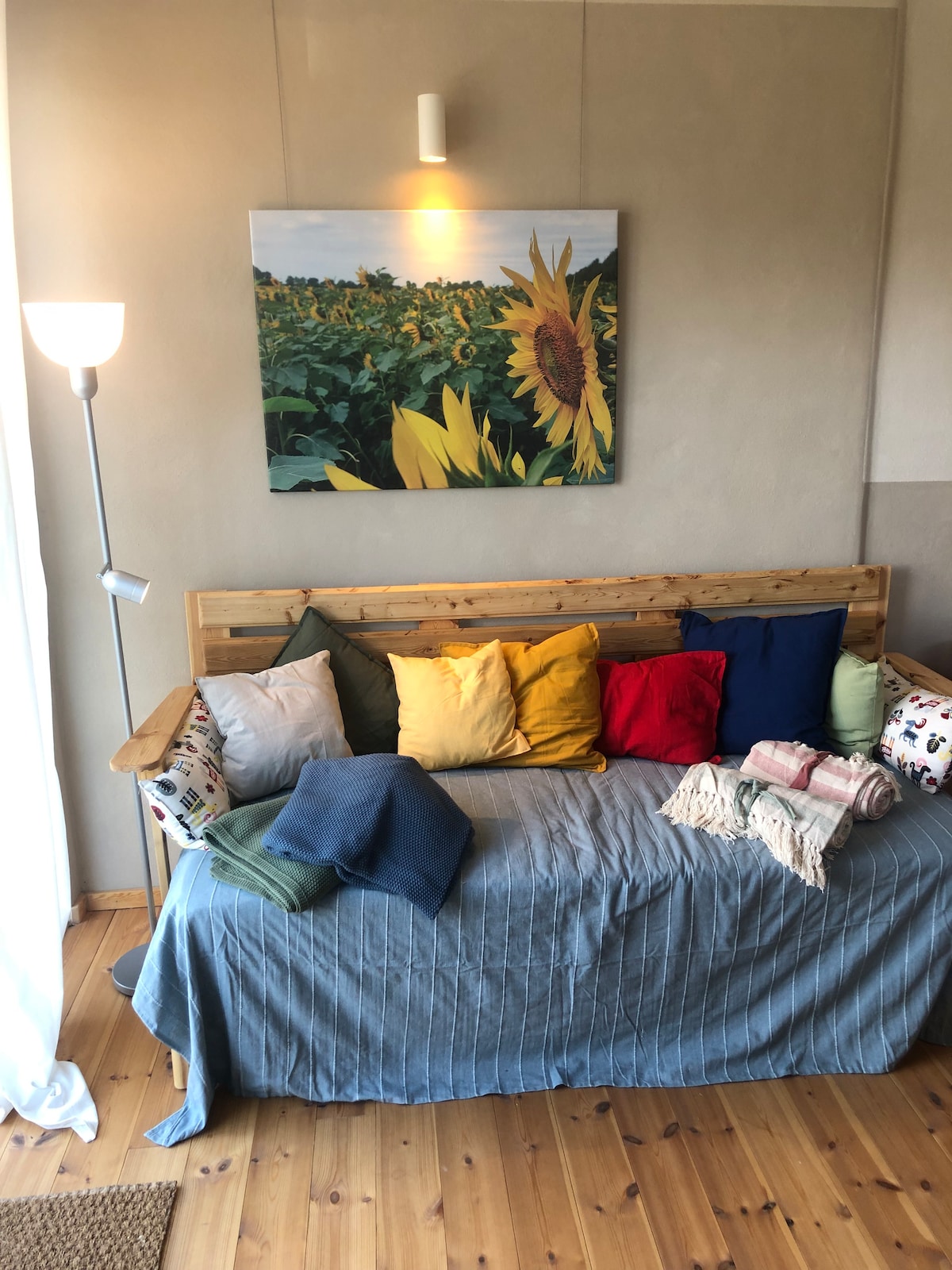 A cozy seating area features a wooden bench adorned with a variety of colorful cushions and blankets. A large artwork of sunflowers hangs on the wall, and soft light from a standing lamp creates a warm ambiance. Natural wood flooring is visible beneath.