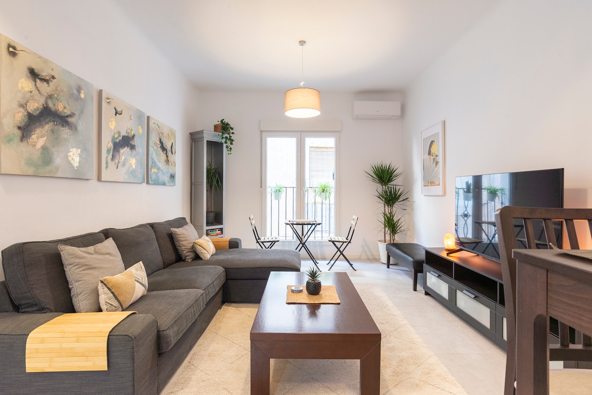A modern living room features a spacious gray sectional sofa adorned with decorative pillows. A wooden coffee table is centered in the room, complemented by a plant and a soft light fixture. High windows open onto balconies, providing natural light. A Smart TV is positioned against the wall.