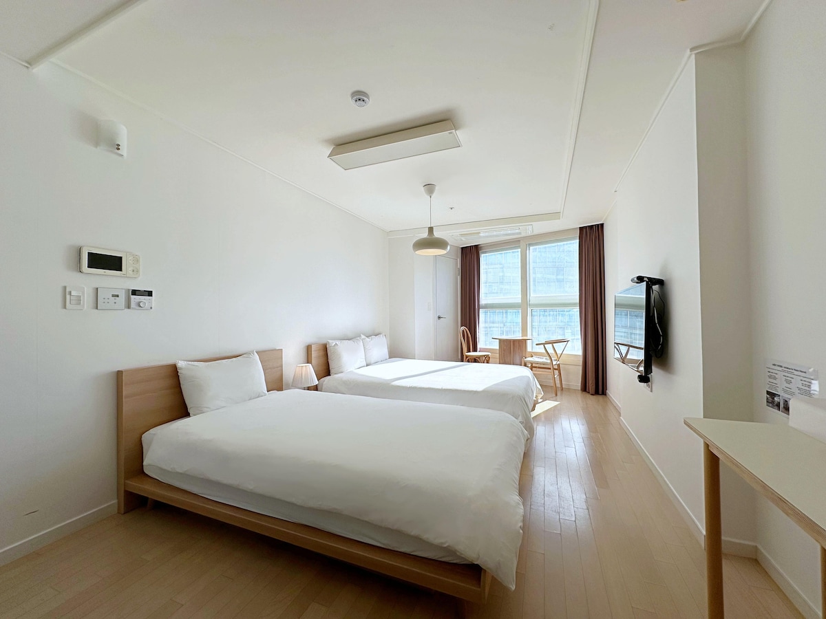 The room features two spacious beds with white bedding, a wooden table, and a chair against the window. Natural light illuminates the space, accentuating the light wooden floors and simple decor. A wall-mounted TV is visible, adding to the room's functionality.
