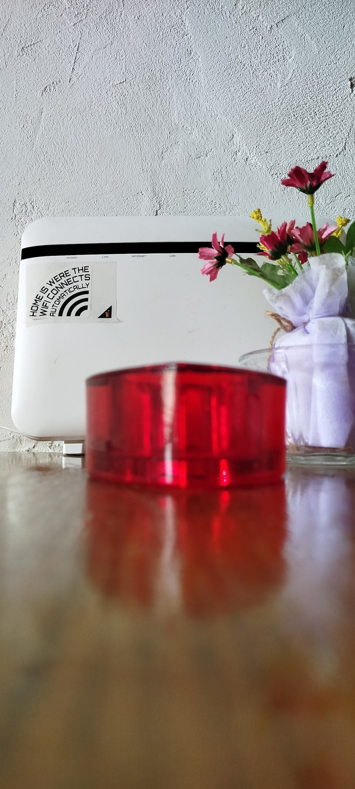 A close-up perspective features a bright red decorative object resting on a wooden surface. In the background, a small glass jar holds a bouquet of flowers, adding a touch of color and life to the space, alongside a white appliance marked with a sticker.