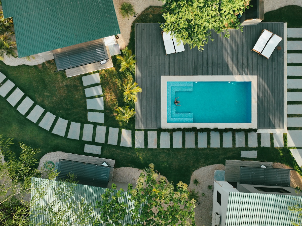 An aerial view captures the arrangement of villas surrounding a rectangular pool, emphasizing the modern wooden deck and lush greenery. The pathway of stone steps leads from the villas to the pool area, offering a serene ambiance amidst the tropical landscape.