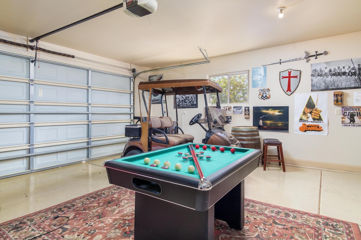 A garage space is highlighted with a bumper pool table at its center, surrounded by a golf cart and various wall decorations, including framed photos and posters. Large garage doors allow for natural light, enhancing the space's ambiance.