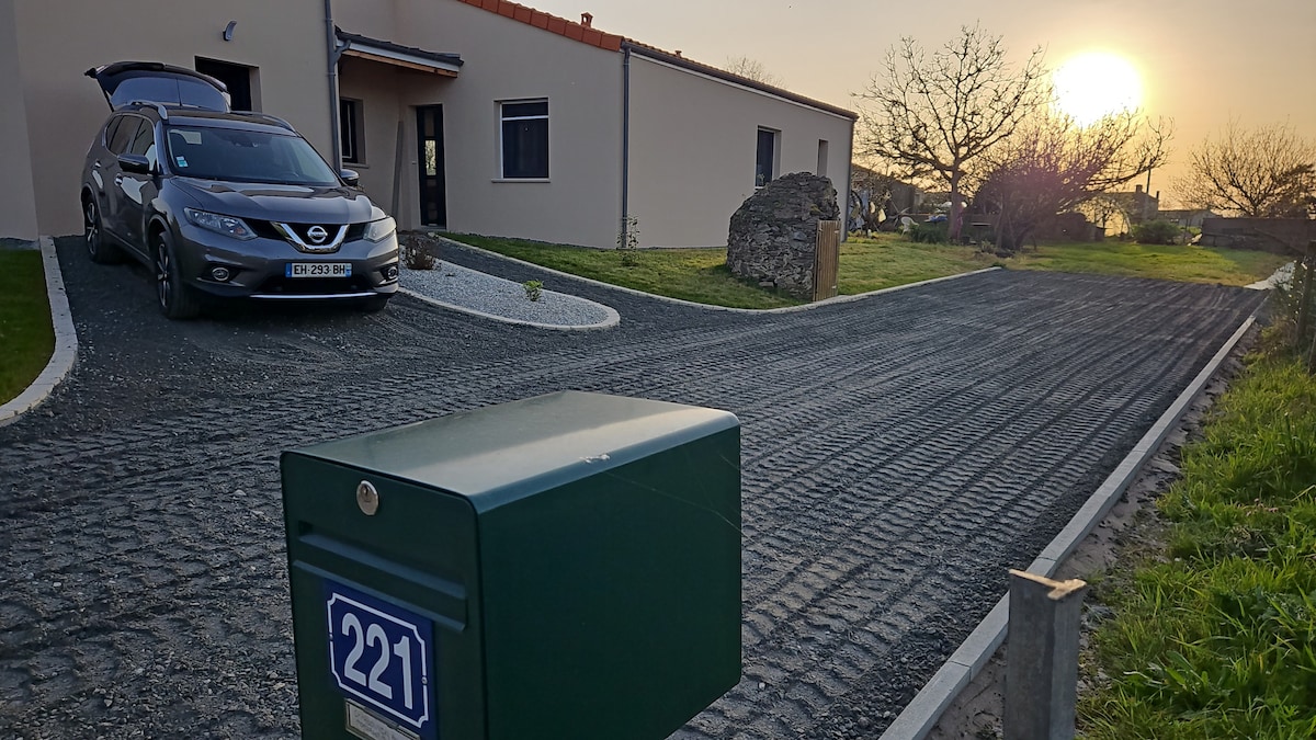 A private entrance is visible, leading to the studio. A vehicle is parked beside the entrance, with a grassy area and a stone feature nearby. The setting sun casts a warm glow over the landscape, accentuating the pathway that guides toward the entrance.