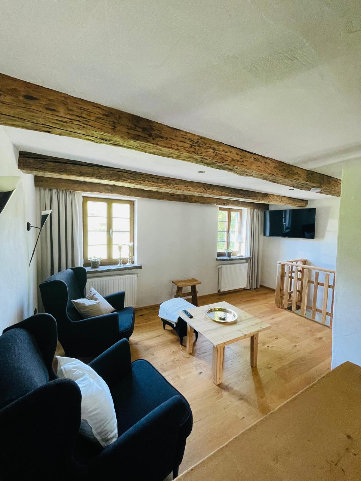 An inviting living area is featured, showcasing exposed wooden beams and large windows allowing natural light to brighten the space. Two dark armchairs are positioned near a light wood coffee table, complemented by a small stool. A flat-screen TV is mounted on the wall.