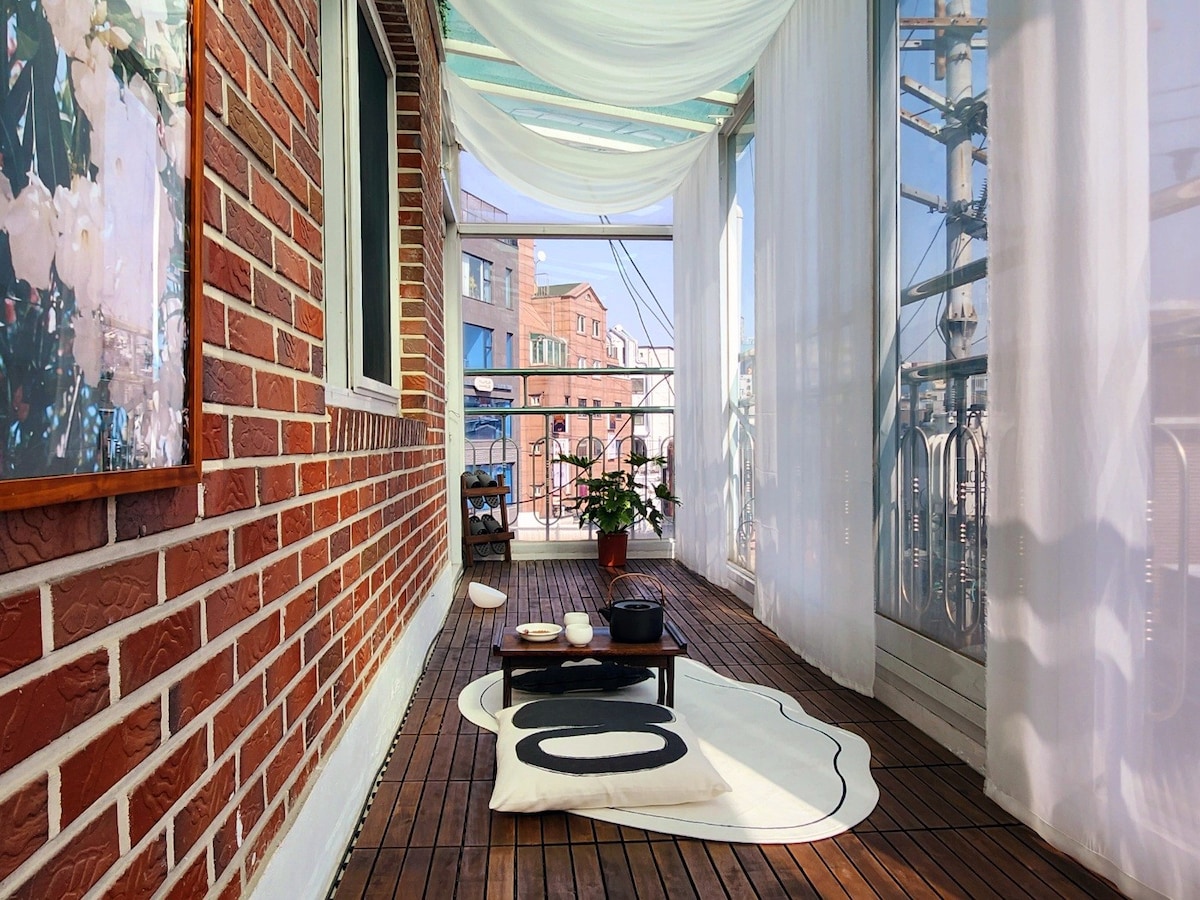 A bright balcony area features wooden flooring adorned with a soft rug. Natural light flows through sheer curtains, creating an airy feel. A small table with items for relaxation is positioned beside a potted plant, offering a serene space for enjoying the outdoors.