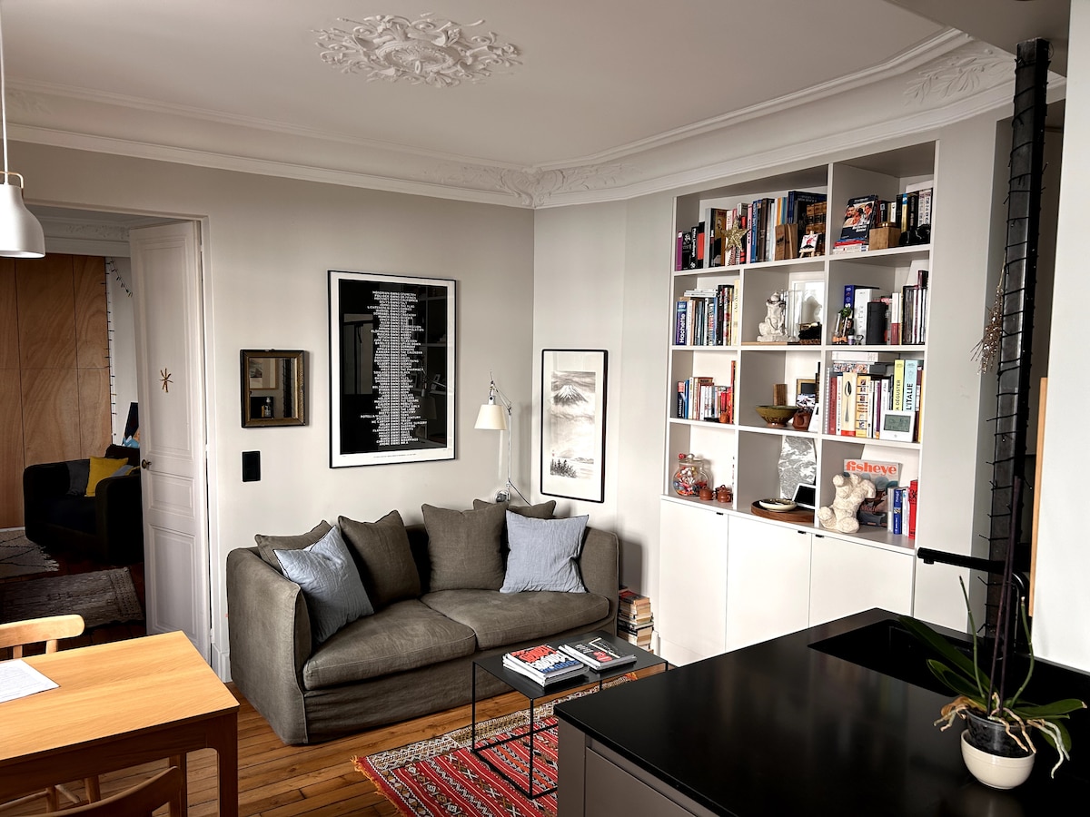 A well-arranged living area features a comfortable gray sofa adorned with blue cushions, positioned beside a bookshelf filled with various books and decorative items. A small dining table is visible, complemented by simple wooden chairs. Natural light brightens the space through a nearby window.
