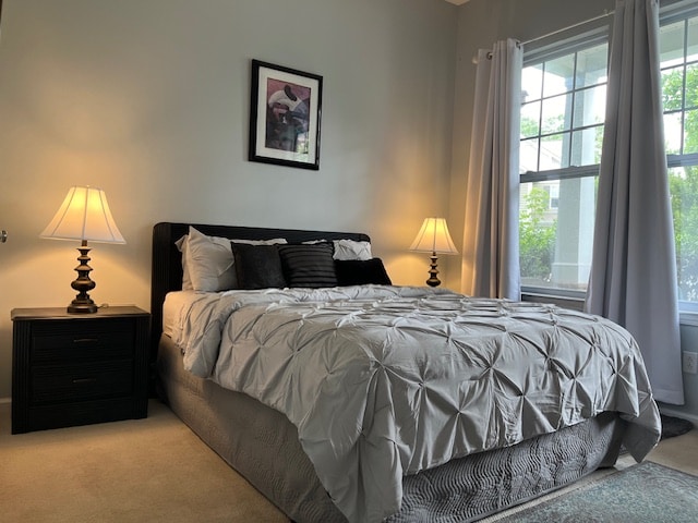 A spacious bedroom features a large bed adorned with a textured gray comforter and multiple pillows. Two bedside lamps provide soft lighting, while large windows allow natural light to illuminate the room. Neutral-colored walls create a calming atmosphere.