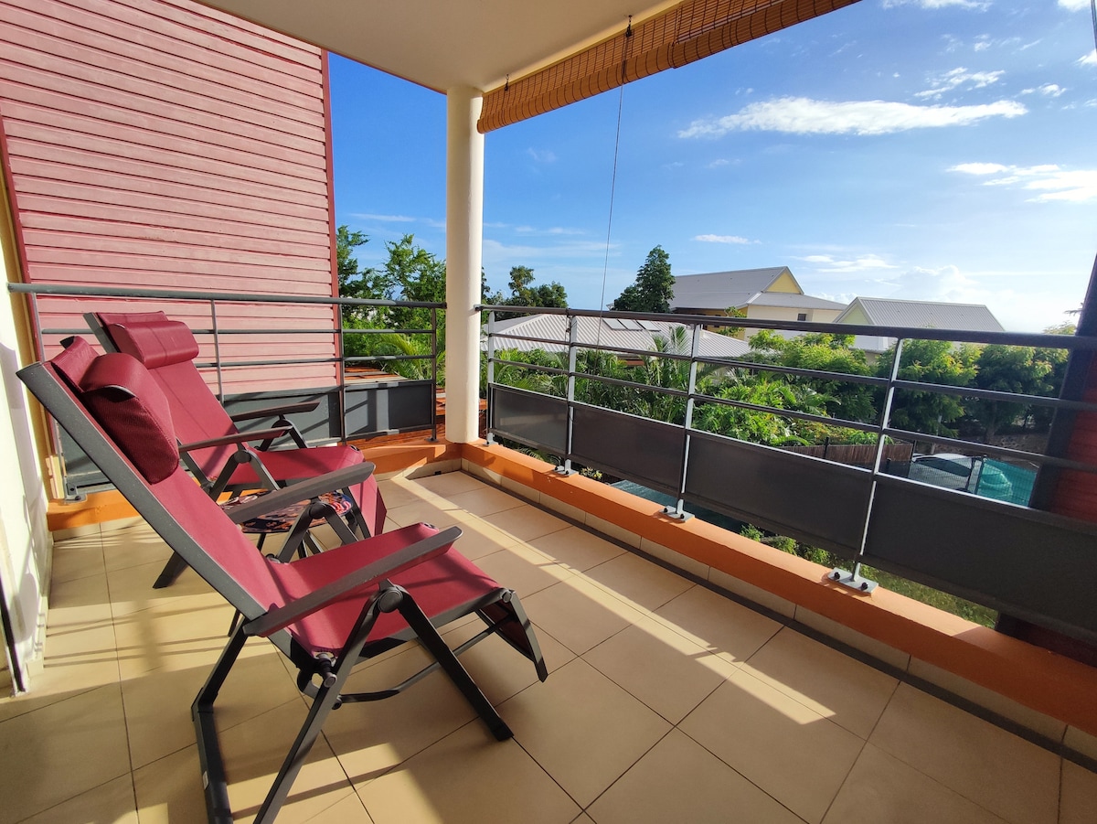 A covered terrace is featured, furnished with two reclining chairs. The glass railing provides a clear view of the surrounding greenery and nearby buildings. Soft sunlight filters through, creating a serene outdoor space for relaxation.