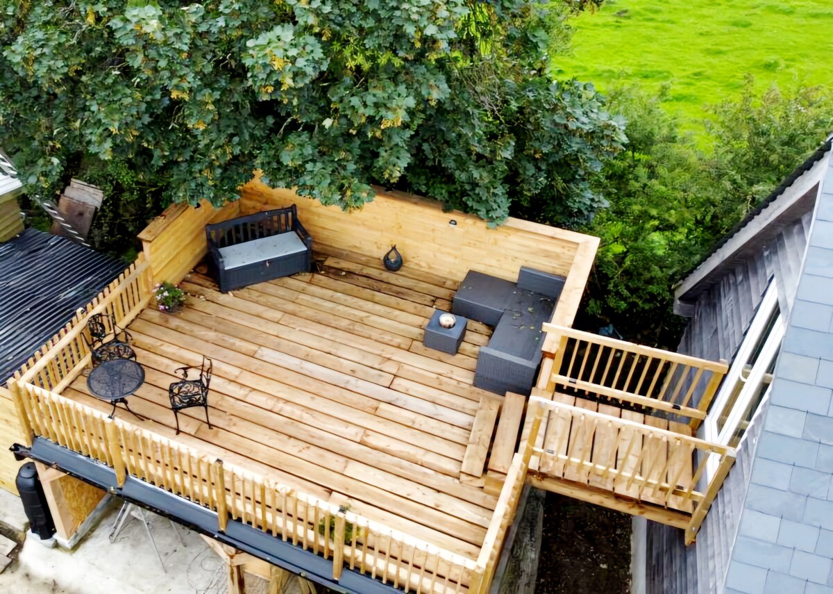A spacious wooden sun terrace is surrounded by green foliage, offering ample seating with modern outdoor furniture. A metal table and chairs are placed for relaxation, while a small decorative feature adds a touch of interest. The terrace connects to the building through a wooden railing.