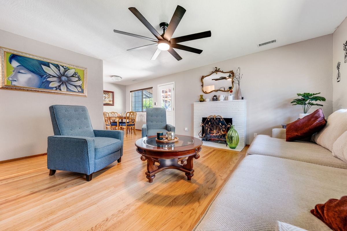 A comfortable living area features a light-colored couch paired with two blue accent chairs. A wooden coffee table sits in the center, surrounded by warm hardwood flooring. Natural light enters through a nearby window, highlighting a decorative fireplace and a glimpse of a dining space in the background.