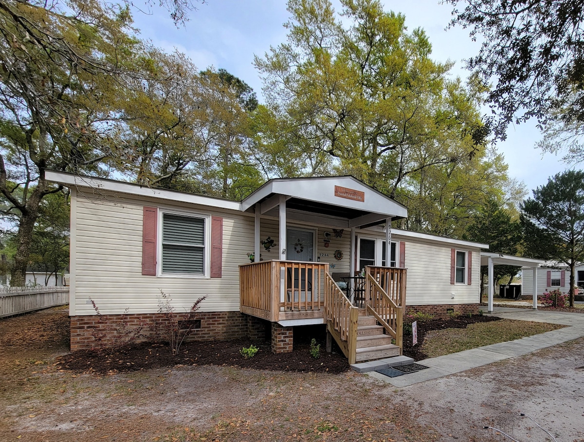 A coastal villa exterior is displayed, featuring a welcoming front porch with steps leading up to the entrance. Surrounding trees offer shade, and a gravel pathway leads to the driveway. The structure has multiple windows, enhancing natural light in the living spaces.