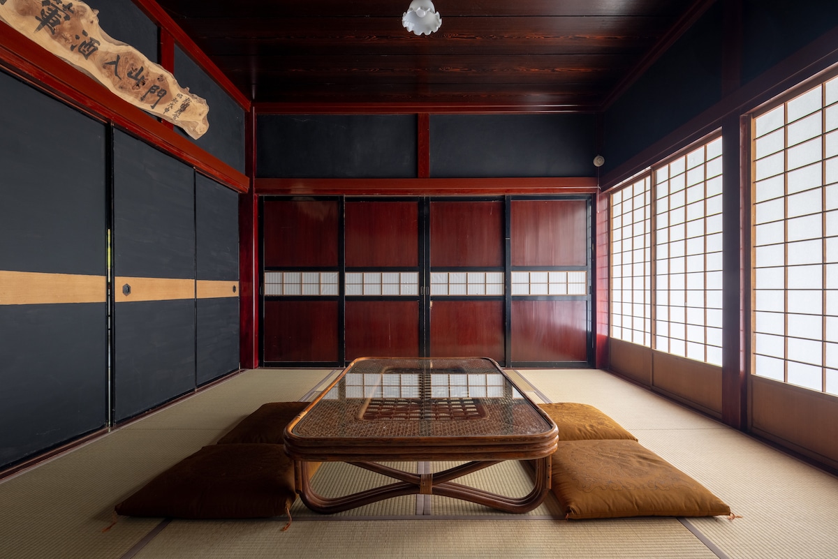 A traditional Japanese room is filled with tatami mat flooring and light-colored wood accents. Sliding shoji screens line the walls, allowing natural light to filter in. A low wooden table is centered among cushions, creating a serene atmosphere ideal for relaxation.