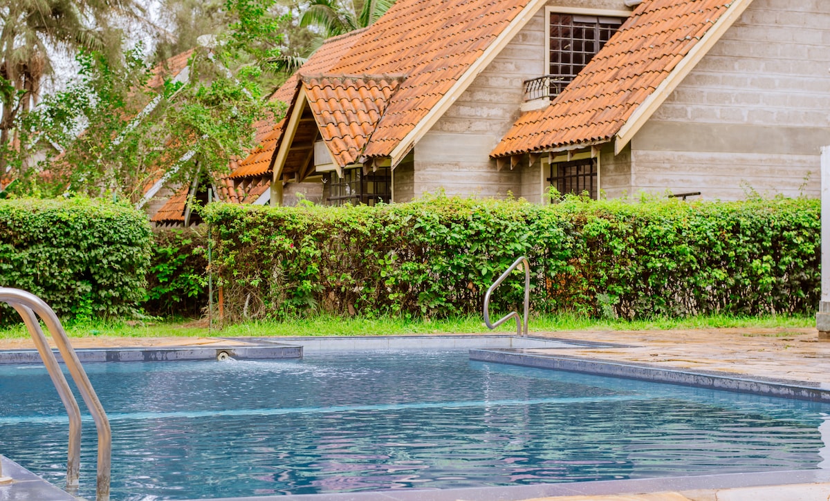 A shared swimming pool is surrounded by lush greenery and a low hedge, providing privacy. The pool area features a smooth surface with steps leading into the water. Beyond the pool, traditional pitched-roof houses are visible, set within the tranquil garden landscape.