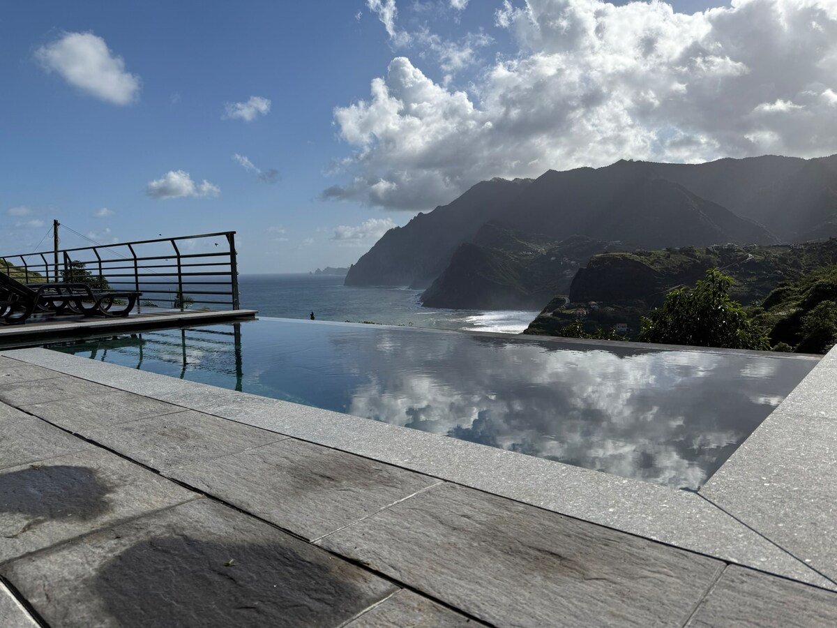 An infinity pool extends towards the horizon, reflecting the sky and mountains. The surrounding patio is designed with stone tiles, and lounge chairs are positioned for relaxation. Coastal views of the sea and lush hills are visible, creating a serene atmosphere.