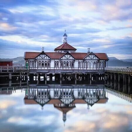 A stunning building is reflected on the calm water, showcasing a design with intricate wooden details and a distinctive tower. The structure features a red-tiled roof and large windows, framed by a tranquil sky above, enhancing the peaceful ambiance of the scene.