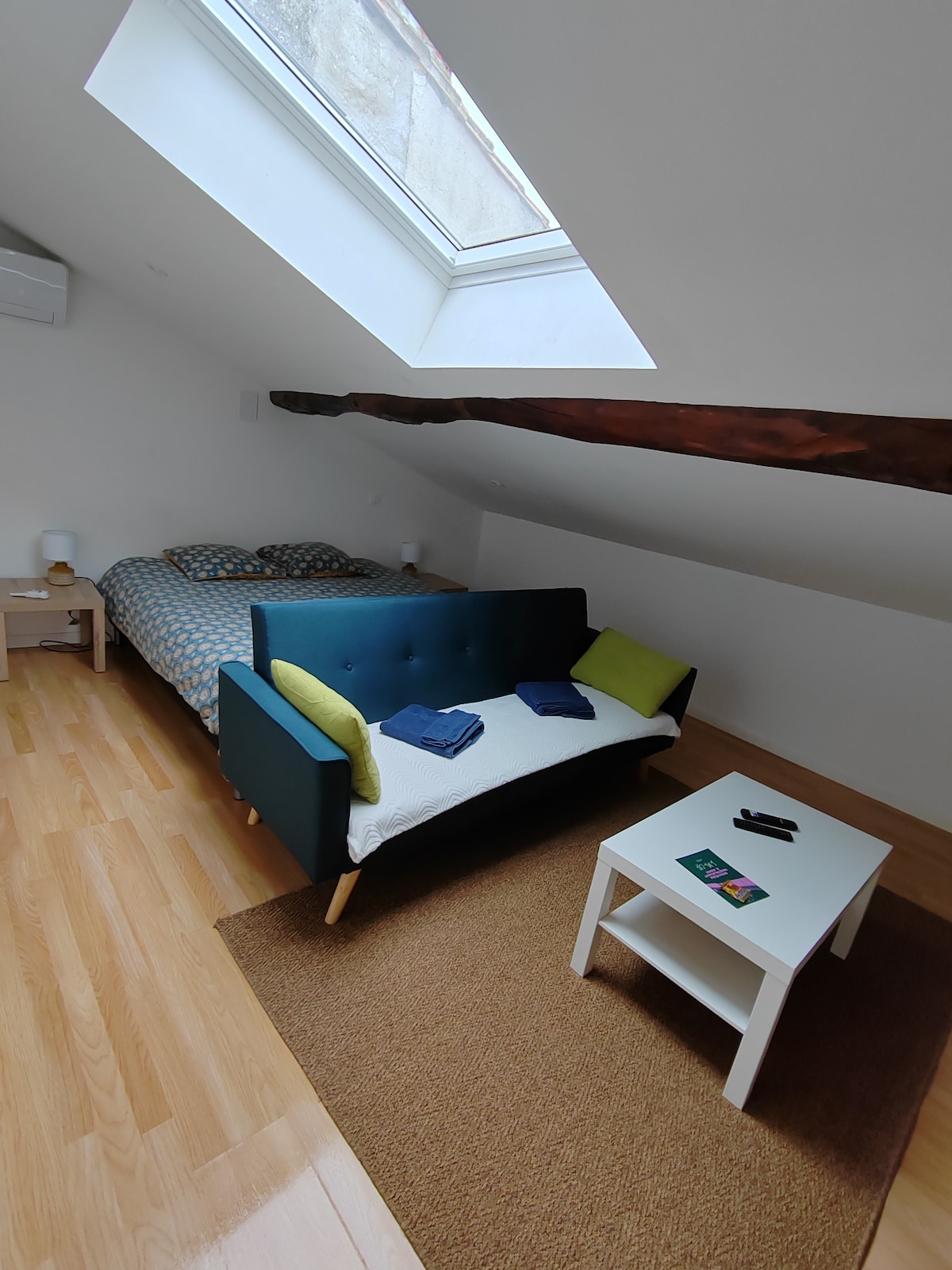 A cozy studio space is illuminated by natural light from a skylight. A spacious bed, covered with a blue checkered blanket, is located at the rear. A small white table sits in front of a dark sofa, adorned with green and blue cushions, creating a functional seating area.