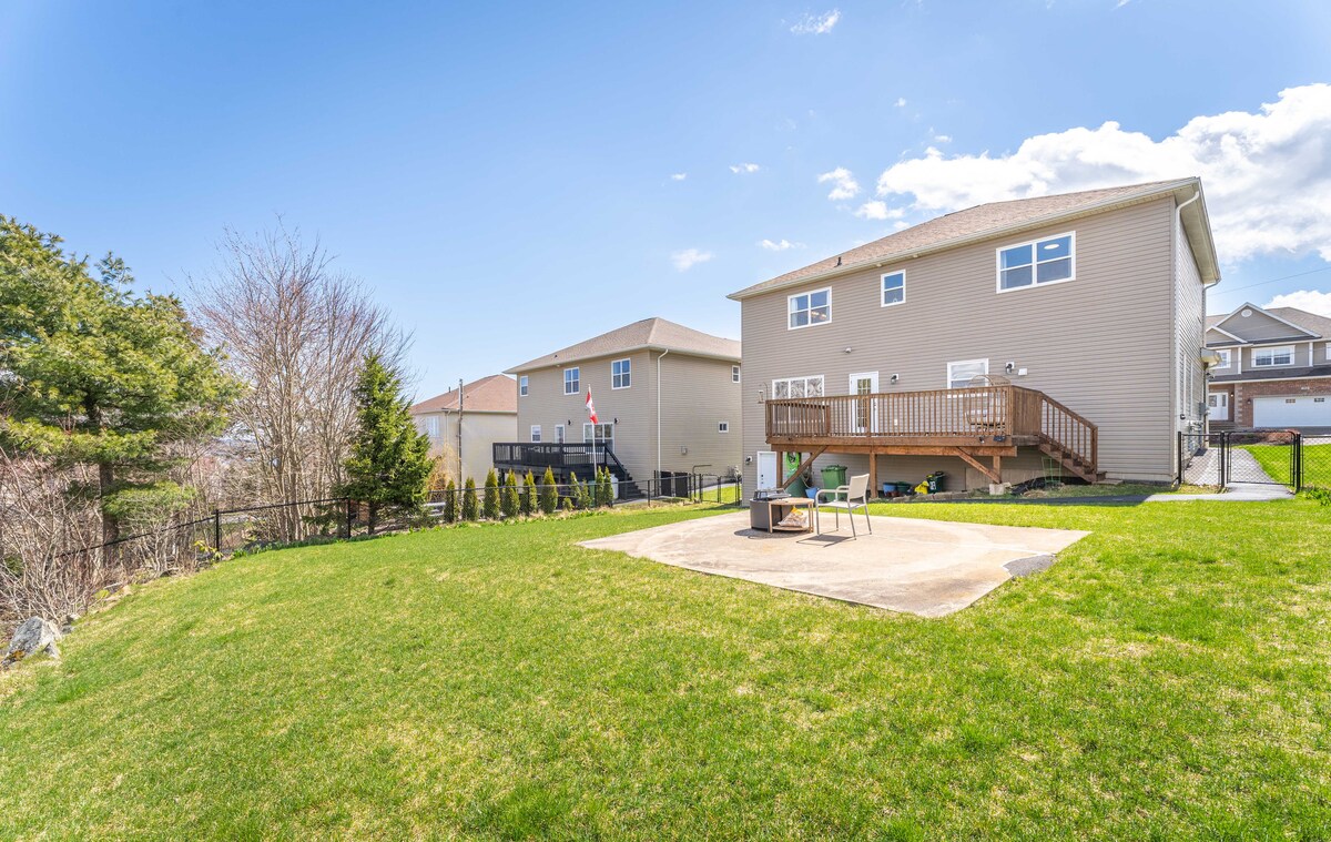 A spacious backyard is highlighted, featuring a well-maintained lawn and a patio area with outdoor seating. The two-story residence is visible in the background, complemented by neighboring houses and trees along the perimeter, all under a clear blue sky.