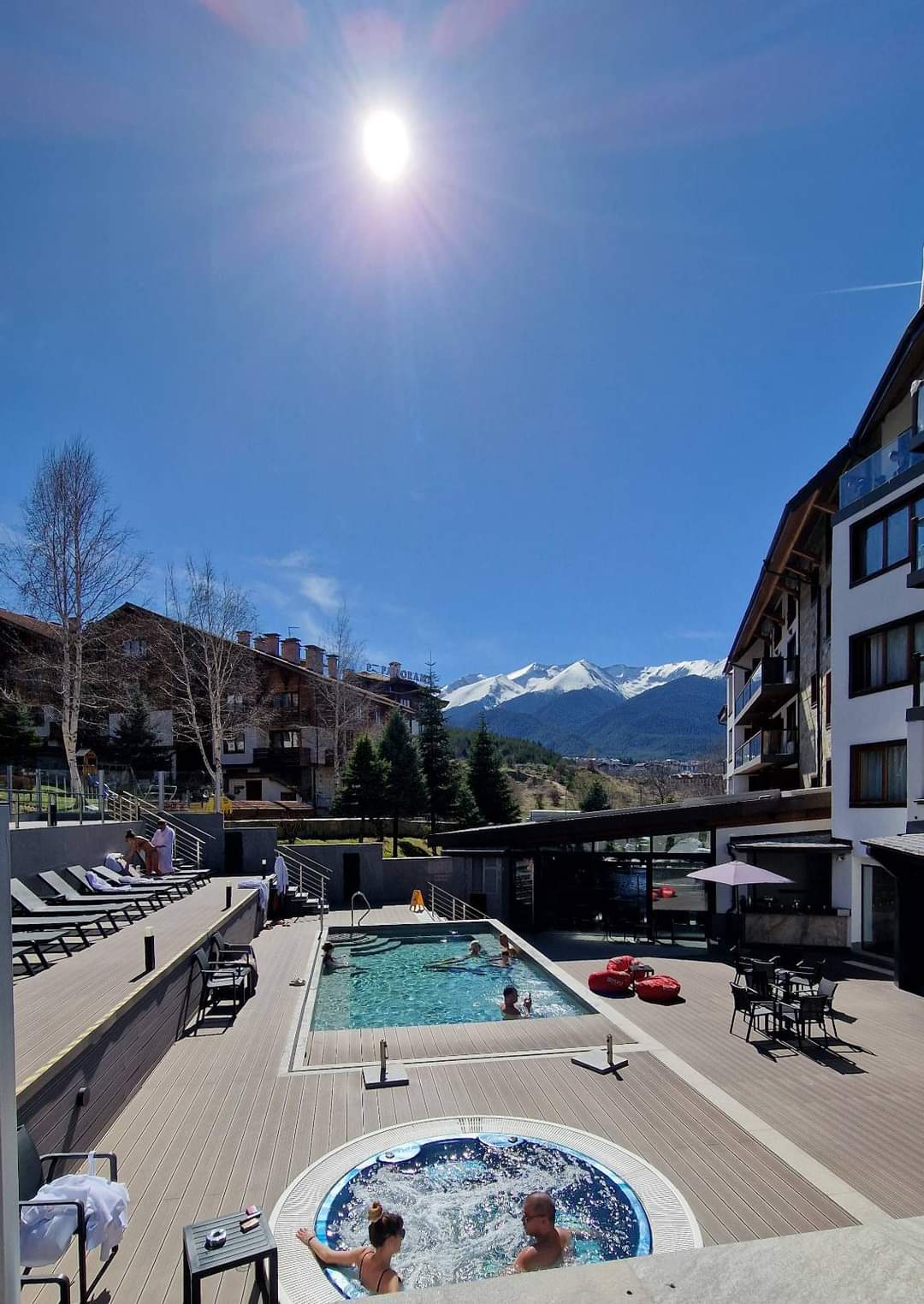 The outdoor area features two swimming pools situated under a clear blue sky. A hot tub is positioned beside the pools, surrounded by lounge chairs. The backdrop is dominated by mountains, enhancing the serene atmosphere of the setting.