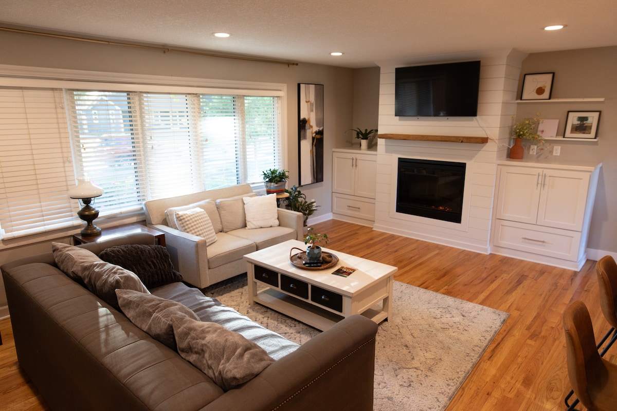 The living area features two comfortable sofas arranged around a central coffee table. A cozy fireplace is framed by a white mantel, and light enters through large windows with blinds. A flat-screen television is mounted on the wall above the mantel, enhancing the modern aesthetic.