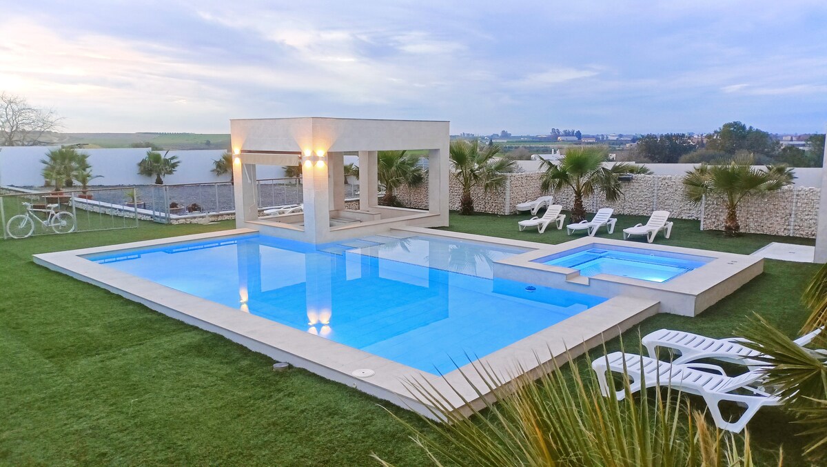 A serene outdoor area features a large swimming pool and a hot tub surrounded by lush greenery. Sun loungers are placed along the poolside, and a shaded pavilion with ambient lighting is visible, providing a perfect spot for relaxation.