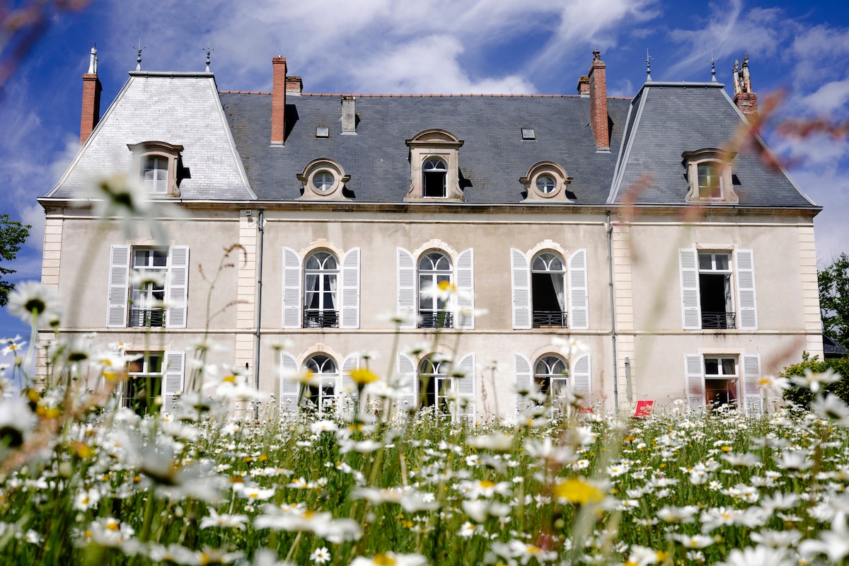 A stately château is surrounded by a vibrant meadow filled with blooming wildflowers. The building features elegant architecture with large arched windows and a slate roof. The peaceful outdoor setting offers a scenic view, highlighting the charm of the historic structure.
