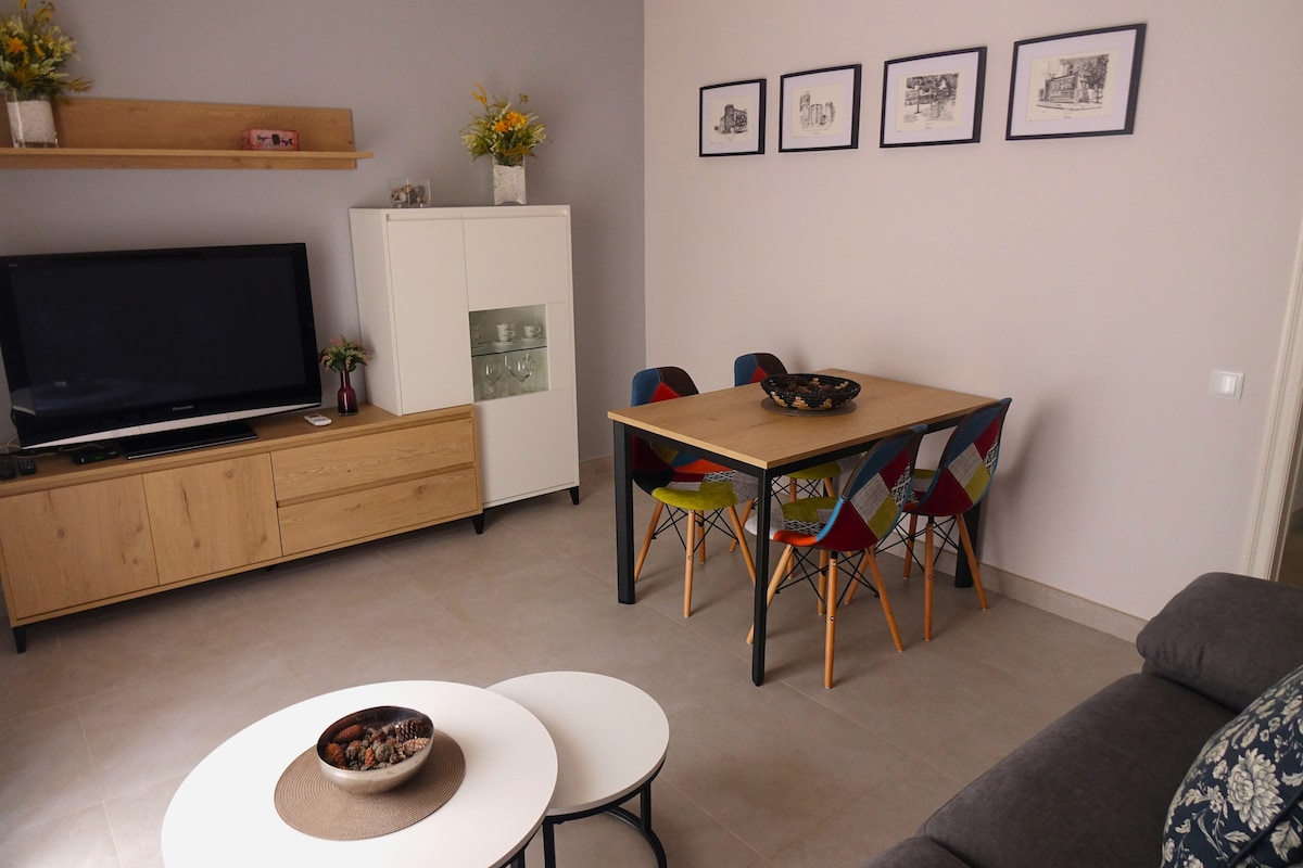 A living area is presented with a soft grey sofa and a stylish coffee table. A dining table with colorful chairs accommodates several guests. A media console houses a television, while framed artwork adorns the walls. Natural light enhances the open and inviting space.