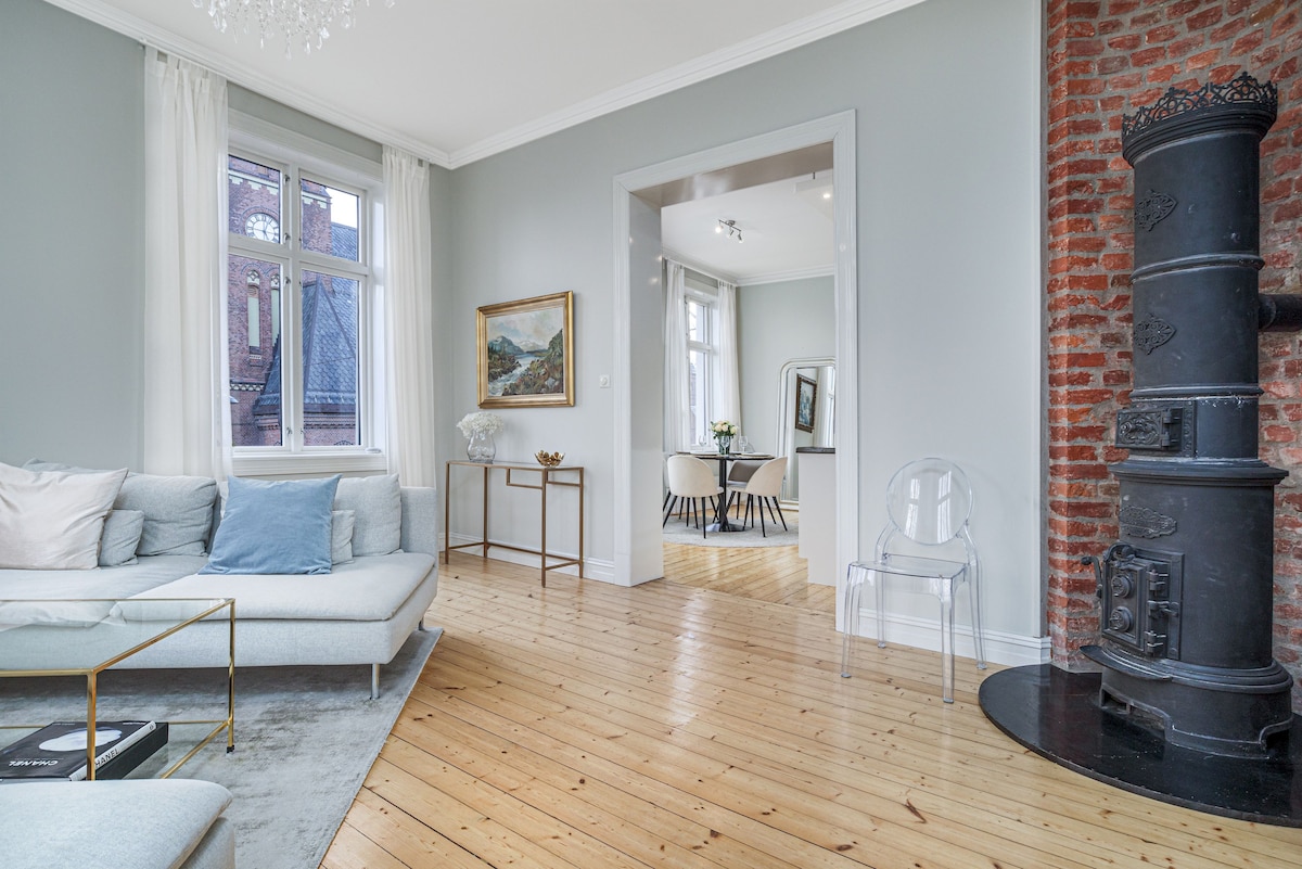 A light-filled living area features a soft, upholstered sofa and a decorative coffee table. The space includes a charming exposed brick chimney, contrasting with the light grey walls. Natural light enters through large windows, illuminating the wooden floor and enhancing the room's welcoming atmosphere.