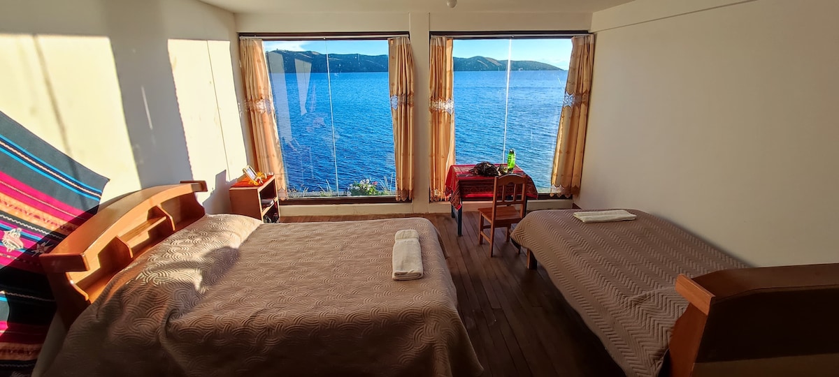 A bright bedroom features large windows offering panoramic views of Lake Titicaca. Two beds are positioned near the windows, while a small table and chairs are set against the wall. Natural light fills the space, highlighting the wooden flooring and soft textiles.