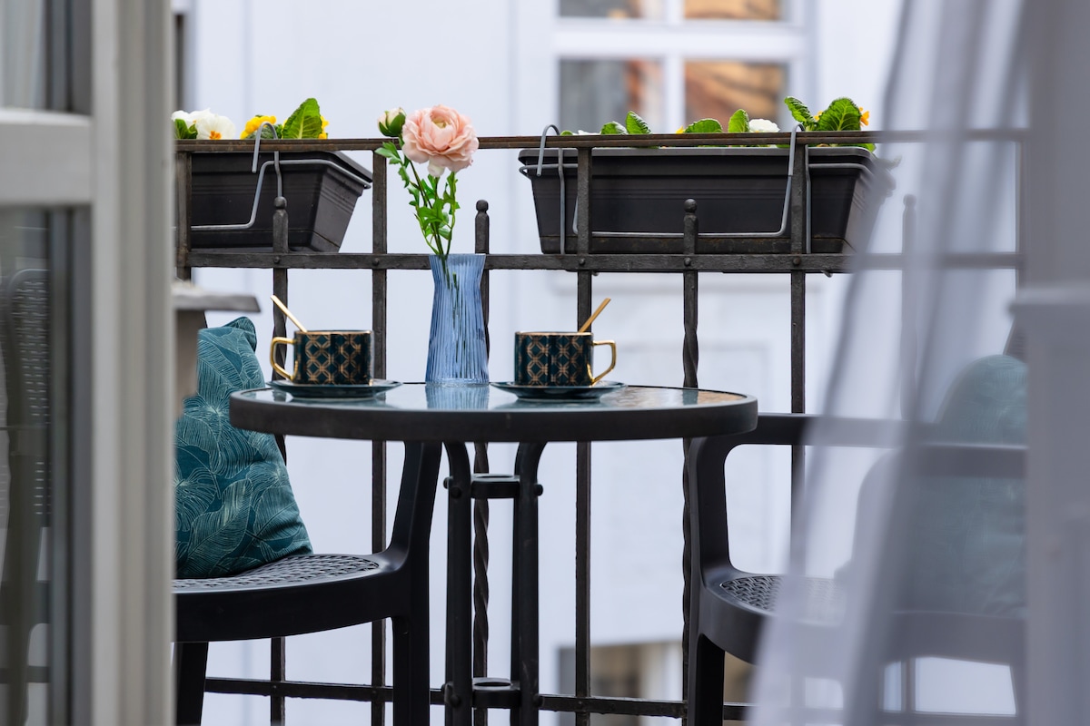 A small balcony features a round table with two black chairs, each adorned with decorative cushions. A blue vase holds a flower, while two mugs are placed on the table amidst lush planters.