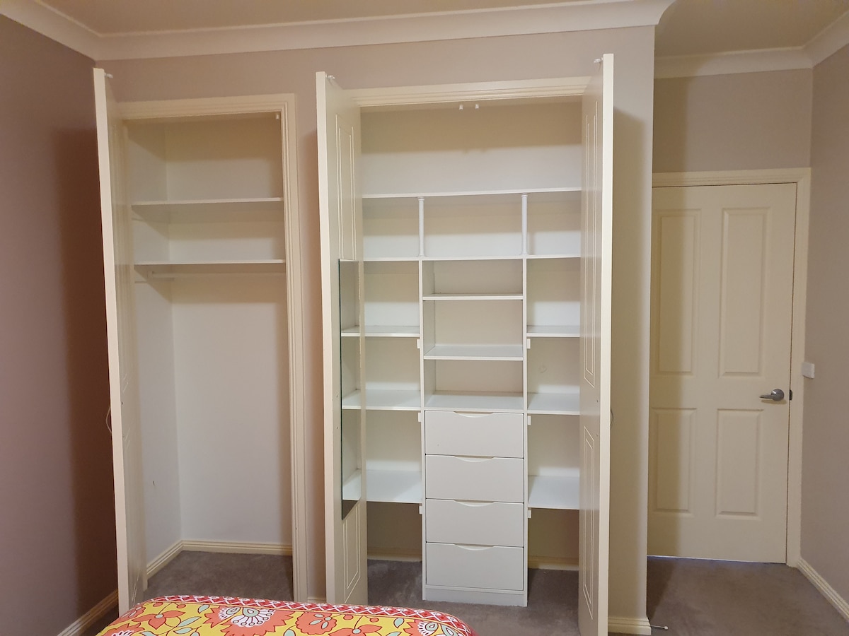 A well-organized wardrobe features open shelving on either side and a central section containing several drawers. The light-colored cabinetry complements the neutral wall tones, while soft carpeting adds to the room's comfort.