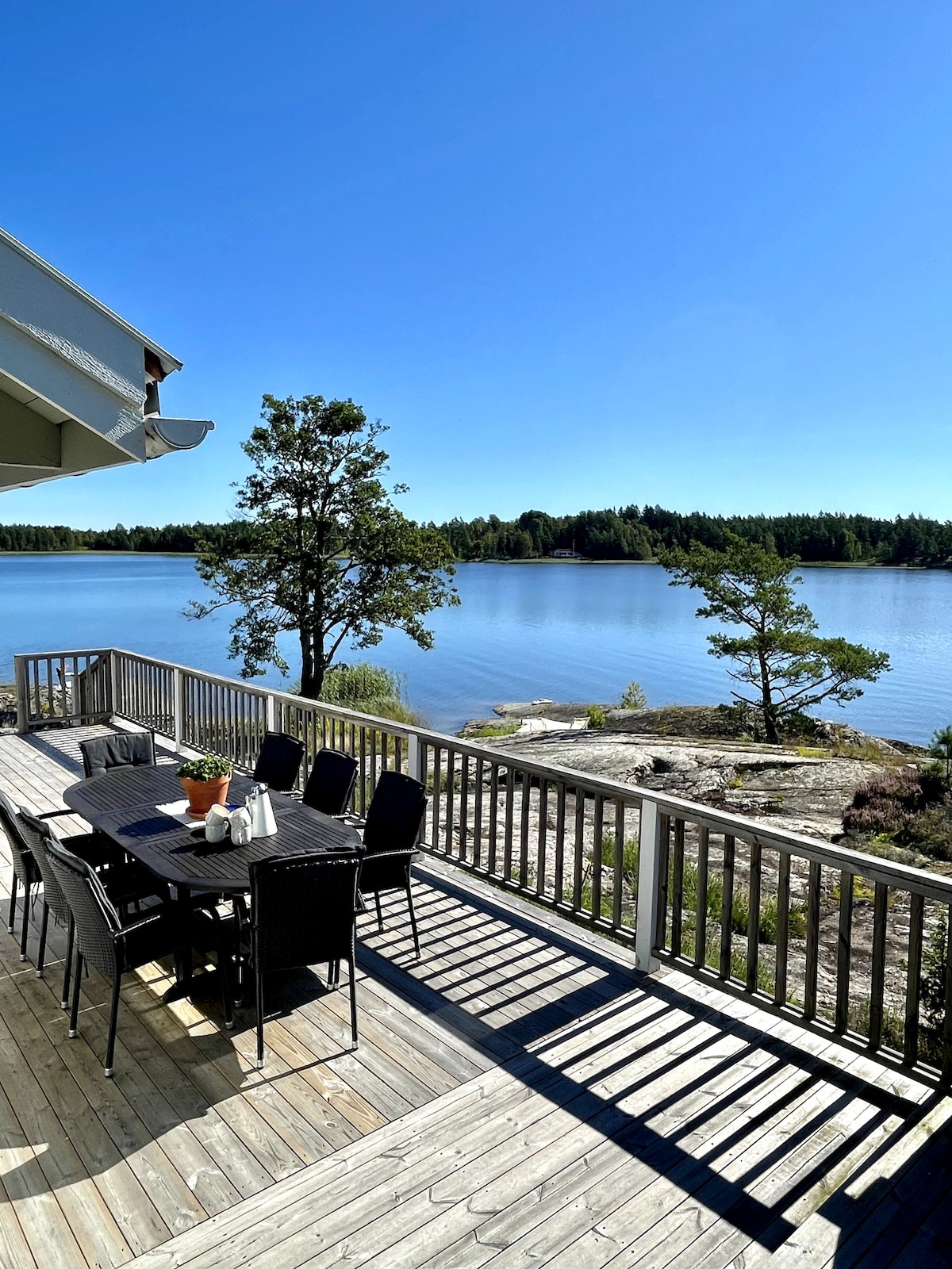 A spacious outdoor deck is shown, equipped with a large dining table surrounded by chairs, offering a waterfront view. Sunlight reflects off the calm water, and trees are visible along the shore, providing a serene natural setting.