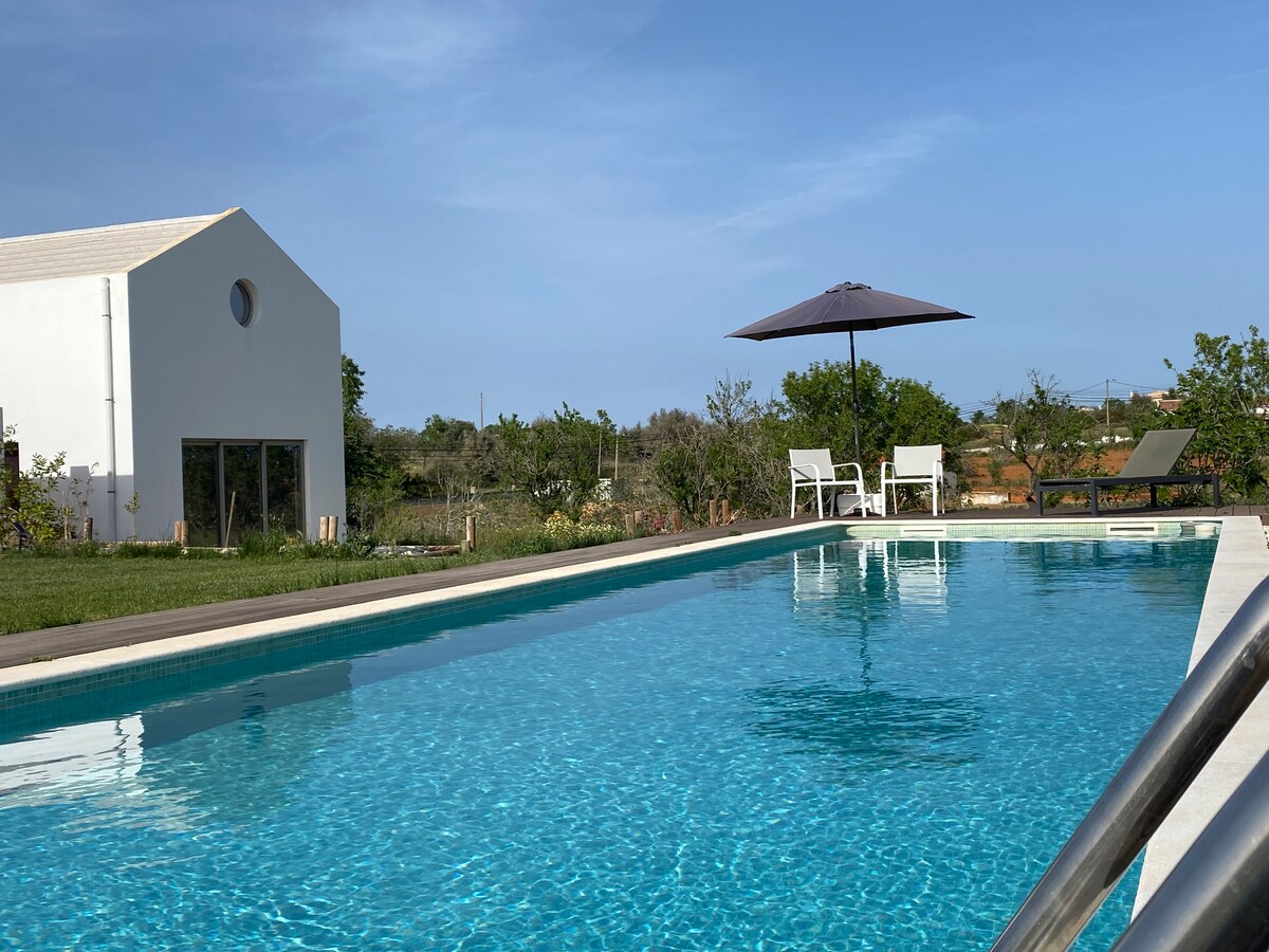 A crystalline swimming pool reflects the sky, surrounded by well-maintained greenery. Comfortable lounge chairs are positioned under a shaded umbrella, offering a relaxing space to enjoy the tranquil surroundings. A modern white structure is visible in the background, enhancing the serene outdoor atmosphere.