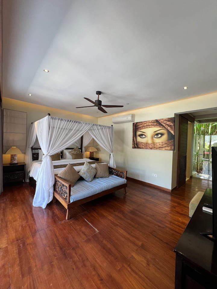 Unique bedroom with a four-poster bed draped in sheer white fabric, complemented by a plush gray sofa.  A large, striking portrait hangs on the wall, adding a touch of drama.