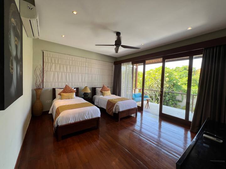 Cozy bedroom with two cozy single beds, wooden flooring, and a private balcony with lush views. Perfect for a peaceful retreat in a luxurious villa.
