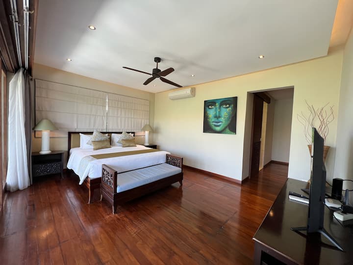 Modern bedroom with a large bed facing floor-to-ceiling windows overlooking a scenic view. Features a ceiling fan and a seating area.