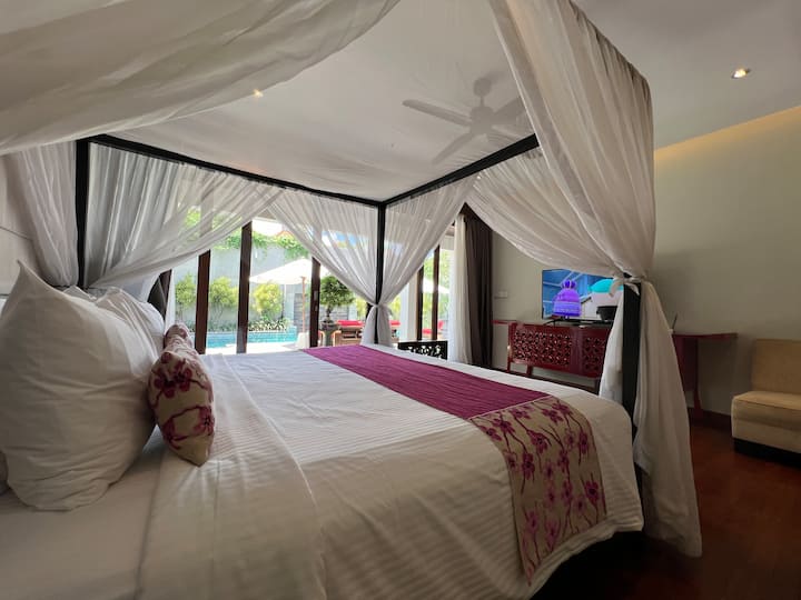 Airy bedroom with a four-poster bed draped in sheer white curtains, creating a dreamy and romantic space.  The room features hardwood floors and opens onto a balcony with scenic views.