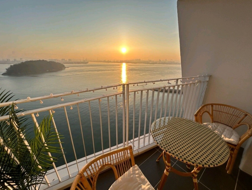 A private terrace offers a serene view of the ocean at sunrise, with soft light reflecting off the water. Two wicker chairs and a glass table are positioned to enjoy the stunning scenery, framed by a railing that enhances the outdoor experience.