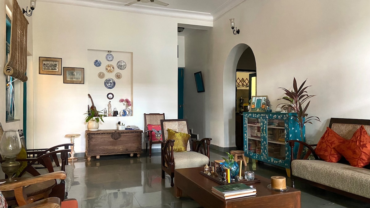 A bright living area features comfortable seating with wooden chairs and a two-seater sofa adorned with colorful cushions. A low coffee table sits centrally, surrounded by decorative items and plants. A vibrant console table adds a pop of color against the neutral walls.