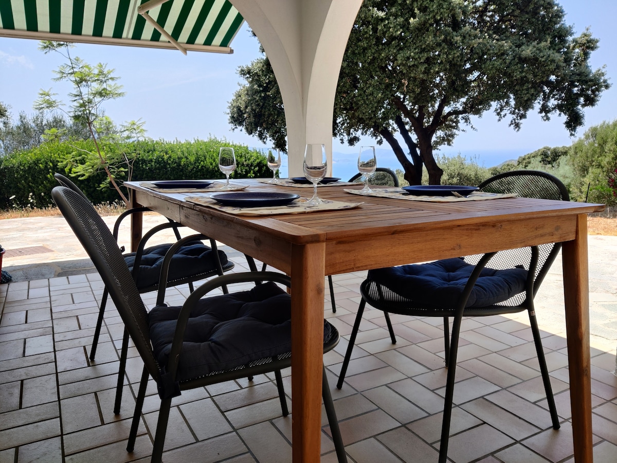 A shaded dining area features a wooden table set for four, with blue placemats and wine glasses. Surrounding chairs exhibit cushions for added comfort. Lush greenery and distant sea views can be seen through the archway, enhancing the relaxed outdoor ambience.