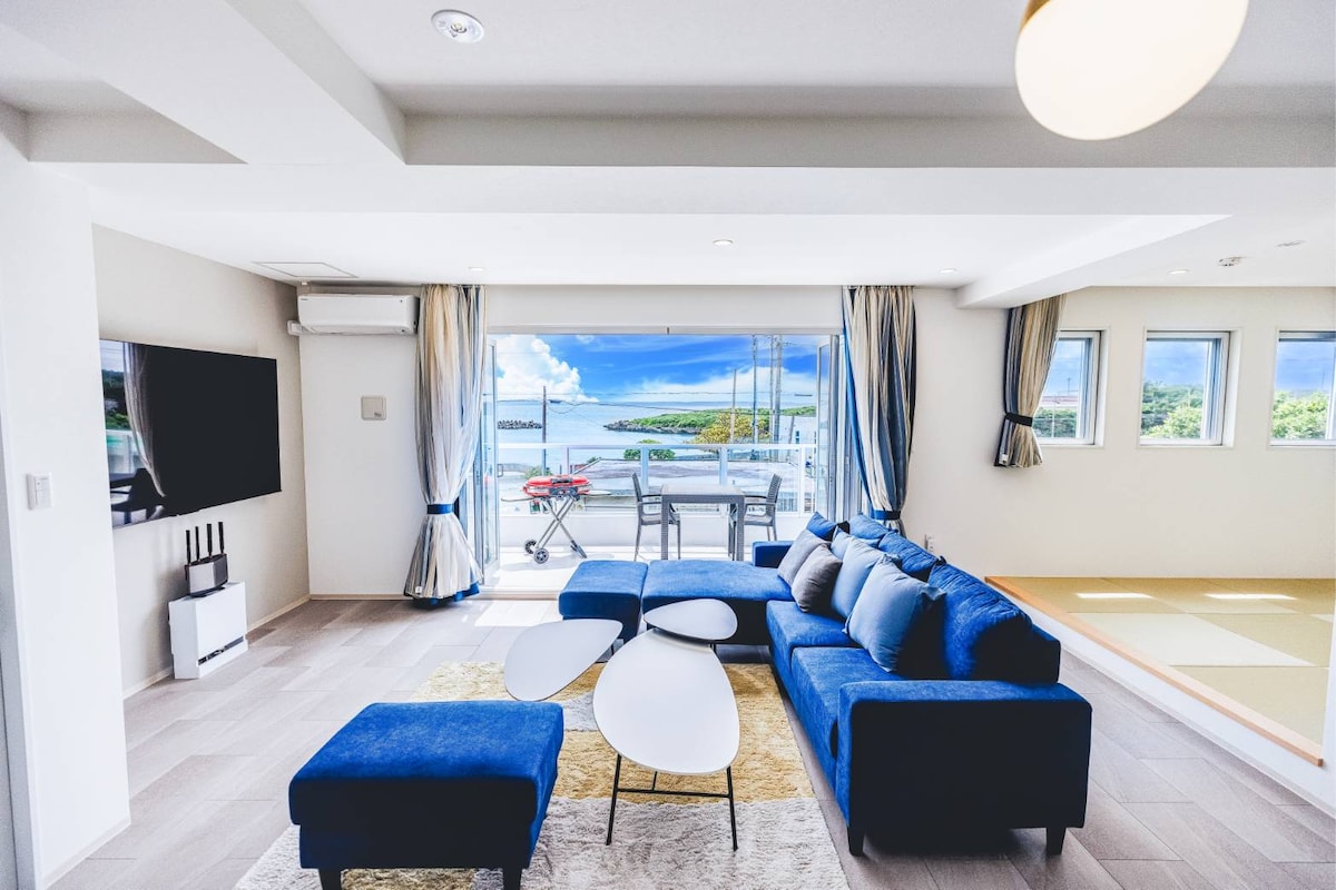 A spacious living area features a blue sectional sofa arranged around a white coffee table. Large glass doors provide a view of the ocean and outdoor seating area, allowing natural light to fill the room. A television is mounted on the wall opposite.