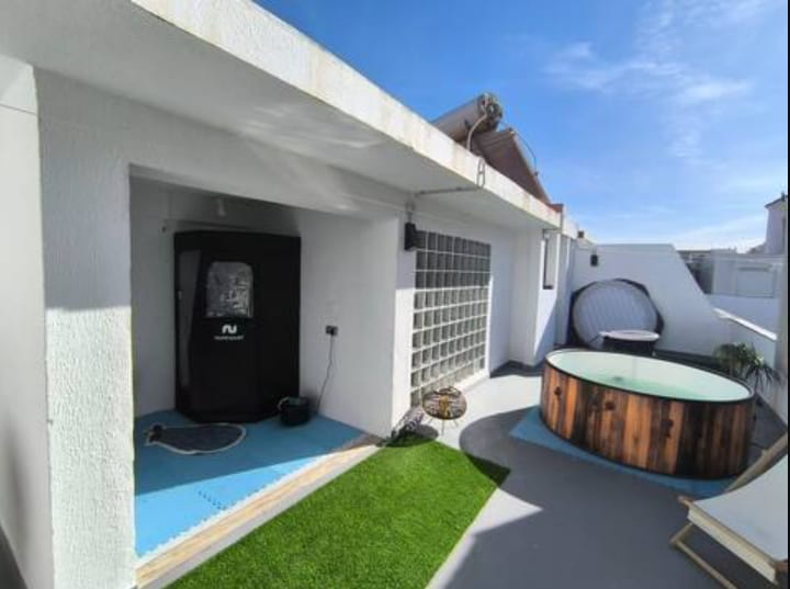 An outdoor terrace features an inviting hot tub surrounded by wooden paneling. A shaded sitting area and a small patch of artificial grass create a relaxed atmosphere. The space is bathed in natural light, with clear blue skies visible above.